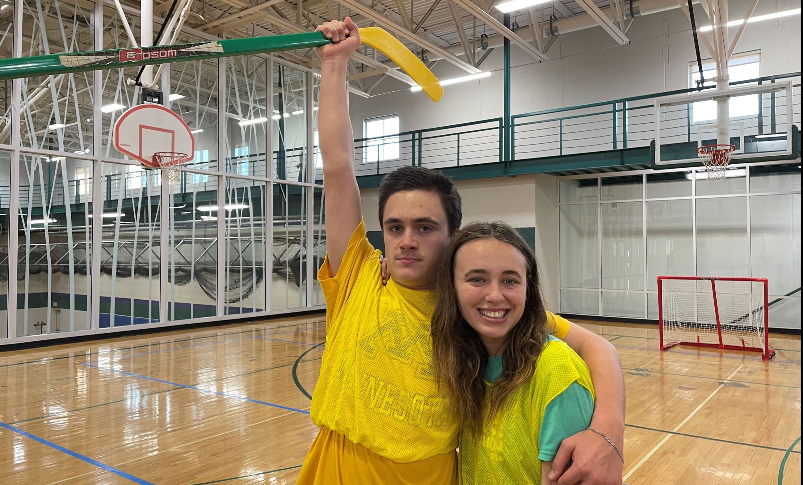 Boy and girl pose for photo inside gym. Boy holds hockey stick above head.